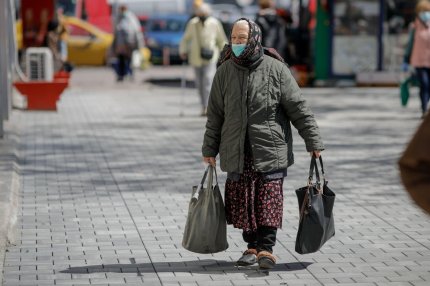 O femeie bătrână se plimbă pe stradă, în România, cu sacoşe pline şi mască de protecţie