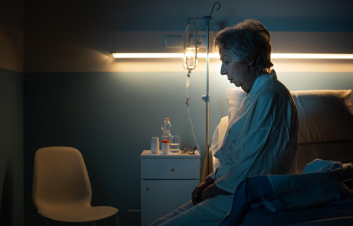 Bătrână pe patul de spital Bătrână îmbrăcată în haine albe, aşezată pe patul de spital, priveşte în jos