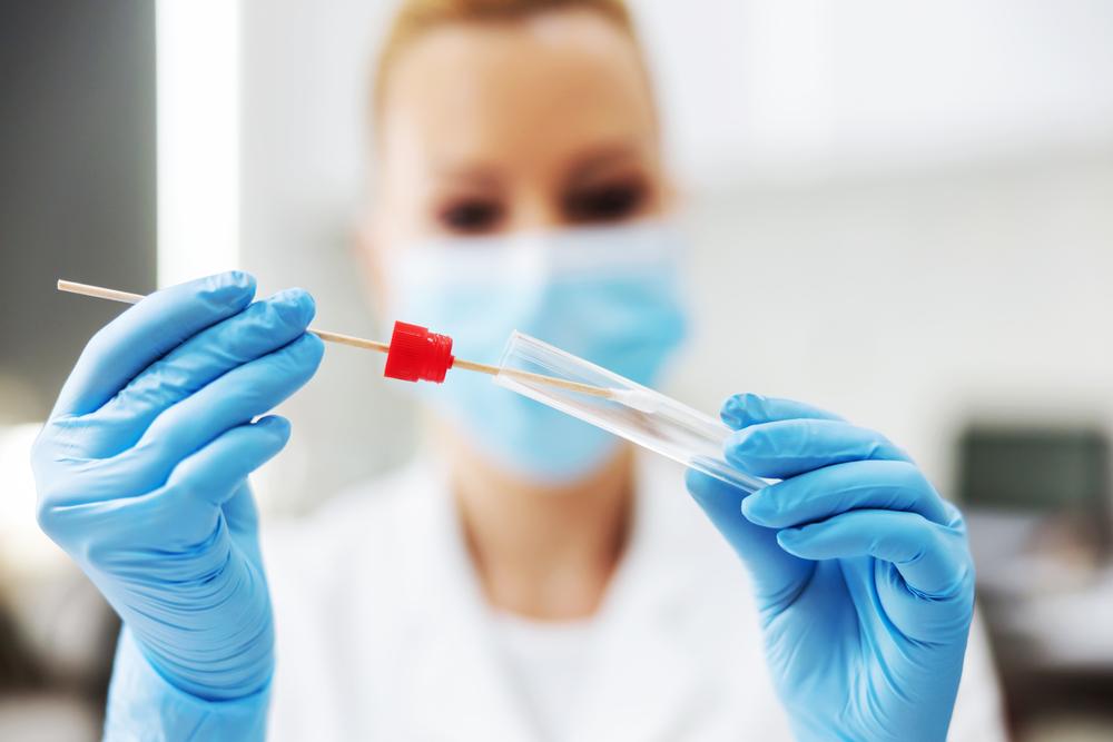 O asistentă de laborator blondă, cu mască şi mânuşi de protecţie ţine un tampon pentru testul Covid-19