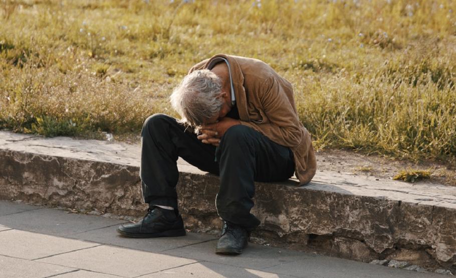 Bărbat fără adăpost Bărbat cu păr cărunt şi geacă maronie, aşezat pe o bordură, cu mâinile împreunate la cap