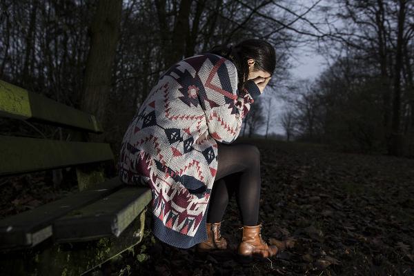 O tânără femeie cu un pulover lung, așezată pe o bancă într-o pădure, în amurg. Ea își acoperă ochii în timp ce plânge