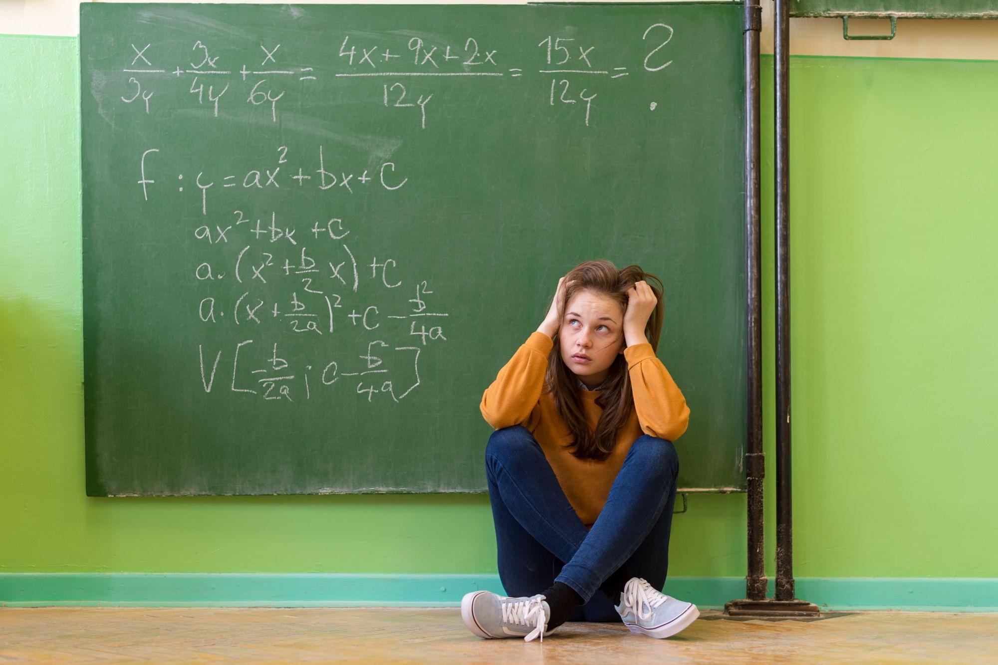 O fată stă pe gânduri cu spatele la o tablă pe care sunt scrise câteva ecuații de matematică