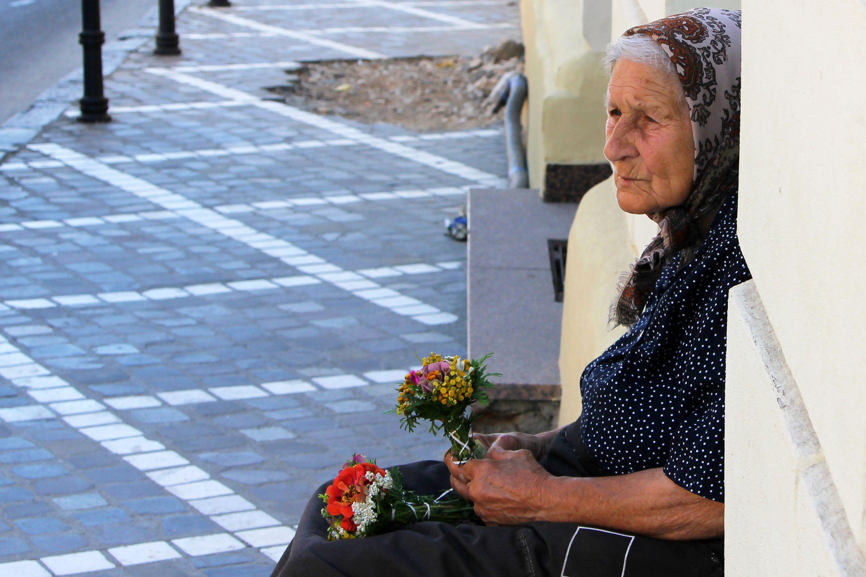 O femeie bătrână stă pe stradă cu două buchete de floricele în mână
