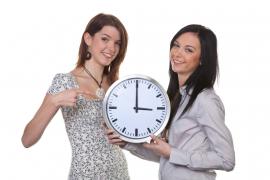 Două fete prezintă un ceas pe care e afișată ora 3, în contextul trecerii de la ora de vară, la ora standard