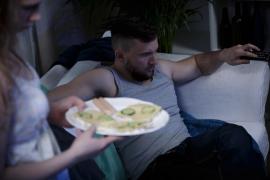 Bărbat pe canapea cu telecomanda în mână, femeie cu mâncare pe o farfurie albă