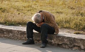 Bărbat cu păr cărunt şi geacă maronie, aşezat pe o bordură, cu mâinile împreunate la cap