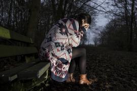 O tânără femeie cu un pulover lung, așezată pe o bancă într-o pădure, în amurg. Ea își acoperă ochii în timp ce plânge