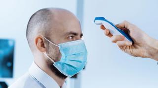 Unui bărbat cu chelie şi barbă, purtând mască de protecţie, i se ia temperatura