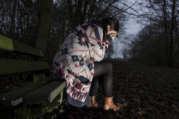 O tânără femeie cu un pulover lung, așezată pe o bancă într-o pădure, în amurg. Ea își acoperă ochii în timp ce plânge