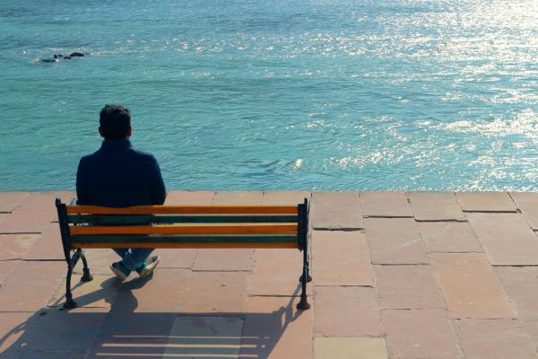 Bărbat singur pe bancă în fața unei întinderi de apa liniștită